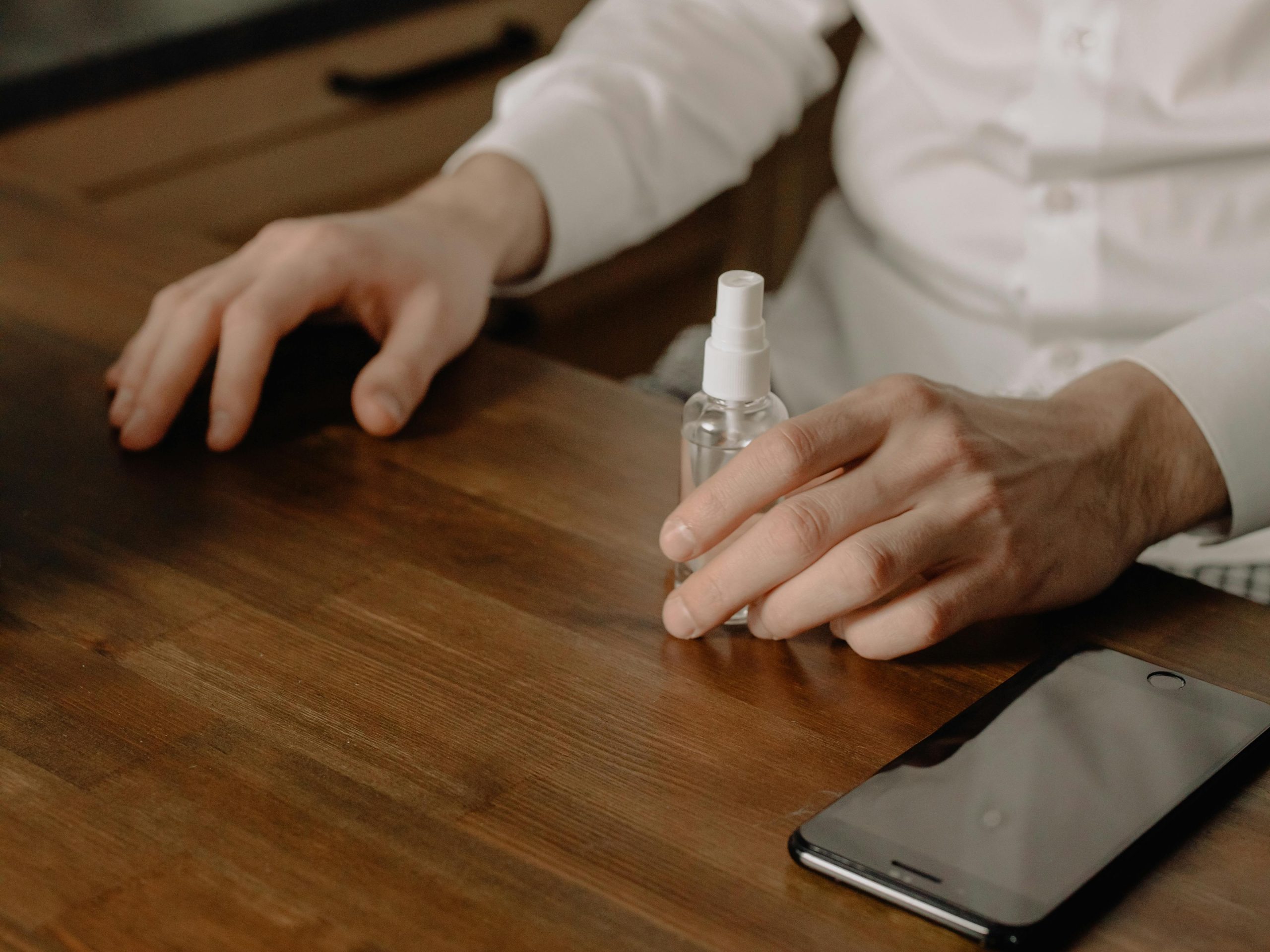 Close-up of hands using sanitizer spray beside a smartphone on a wooden table indoors.